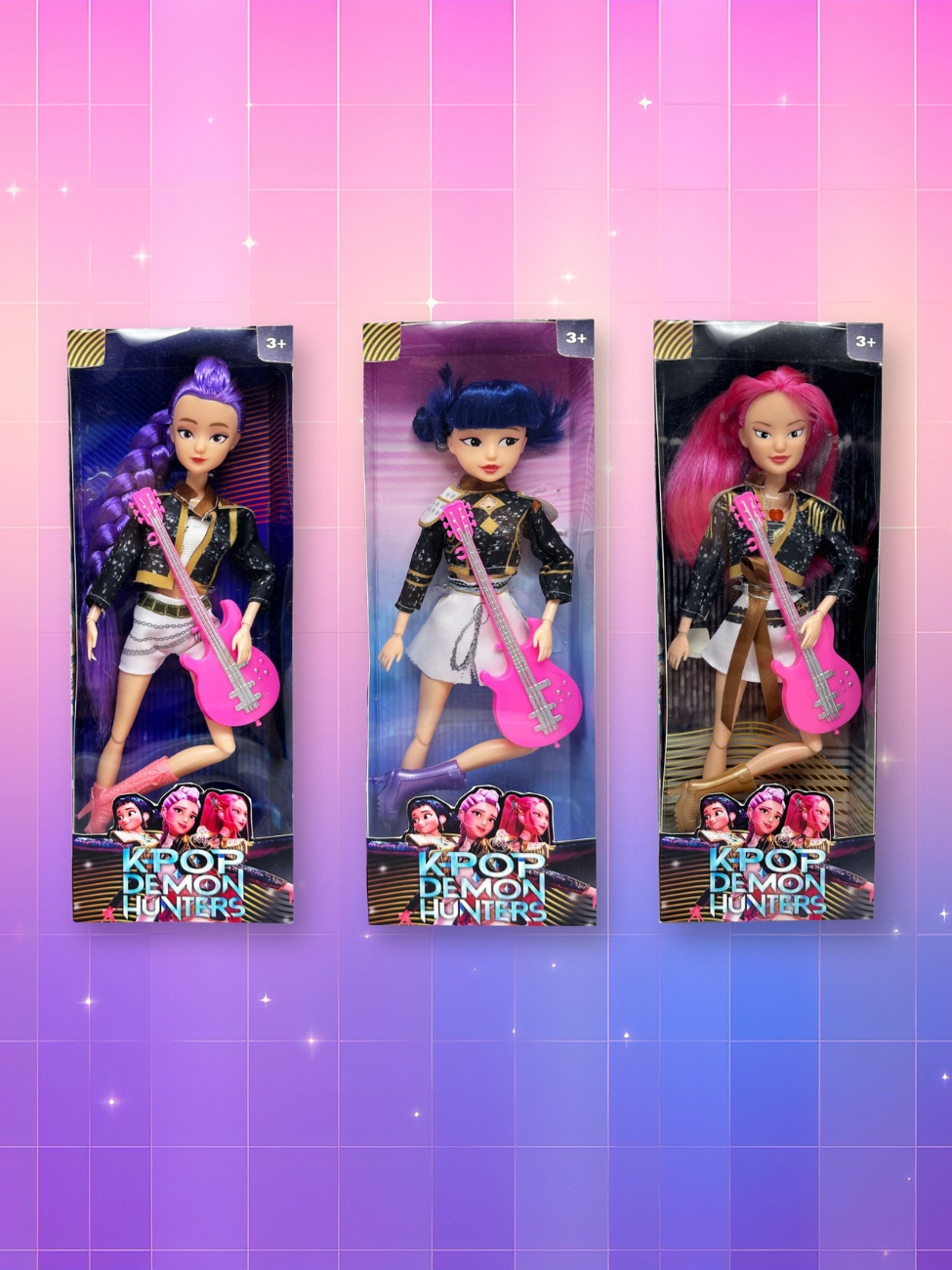 Un lot de 3 poupées Kpop Demon Hunters représentant Rumi, Mira et Zoey, chacune dans sa boîte et avec sa guitare rose, sur un fond artificiel coloré avec un gradient allant du rose au violet.