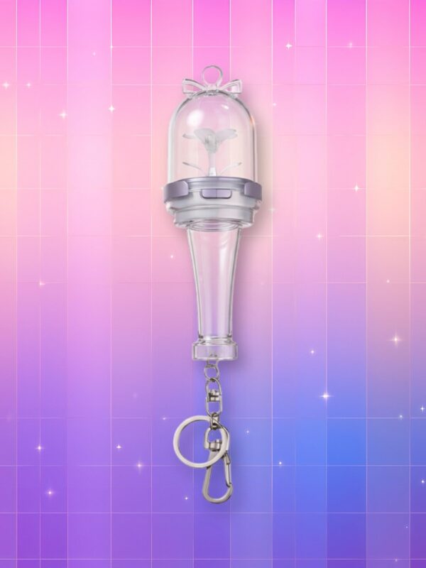 Un Illit lightstick version porte-clés quasiment entièrement en plastique transparent, présentant un manche translucide, un anneau en plastique opaque et un dôme complètement translucide avec une fleur à l'intérieur, sur un fond artificiel coloré avec un gradient allant du rose au violet.