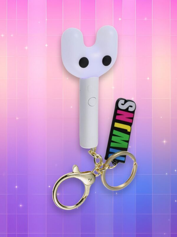 Un lightstick New Jeans Binky Bong version porte-clés entièrement blanc représentant un manche droit dont la tête se sépare en deux branches formant une tête de lapin avec deux billes noires en guise de yeux, accroché à un porte clé coloré avec les initiales du groupe, sur un fond artificiel coloré avec un gradient allant du rose au violet.