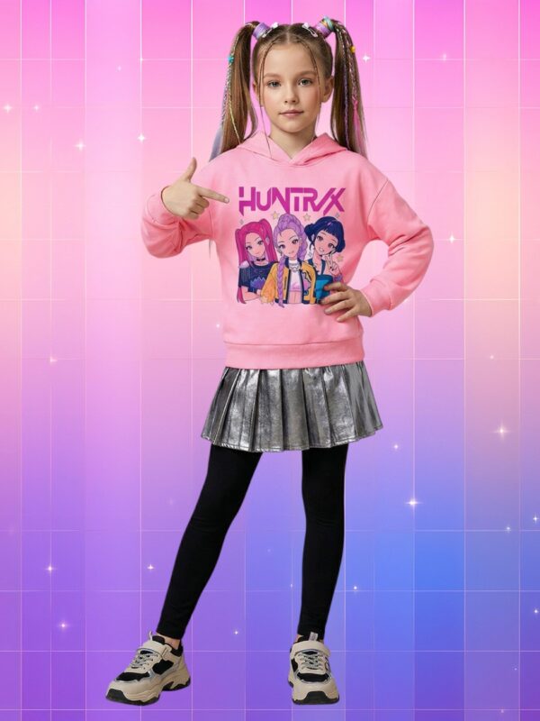 Petite fille en pull Huntrix enfant style K-pop animé pastel rose, montrant fièrement le visuel de trois héroïnes colorées, sur fond rose-violet scintillant.