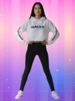 Jeune femme debout en power pose, portant le Pull K-pop Huntrix oreilles style fan girl blanc, crop top avec capuche et manches marquées "DEMON HUNTER", fond rose-violet.