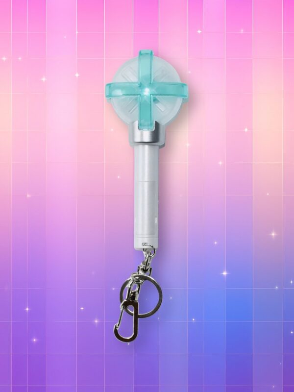 Un lightstick TXT version porte-clés présentant un manche blanc avec trois boutons argentés en forme de croix et un dôme translucide avec une croix verte, sur un fond artificiel coloré avec un gradient allant du rose au violet.