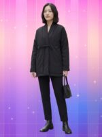 Femme portant une veste kimono femme noire oversize avec pantalon droit, bottines noires et petit sac, sur fond rose-violet à effet brillant et étoilé.
