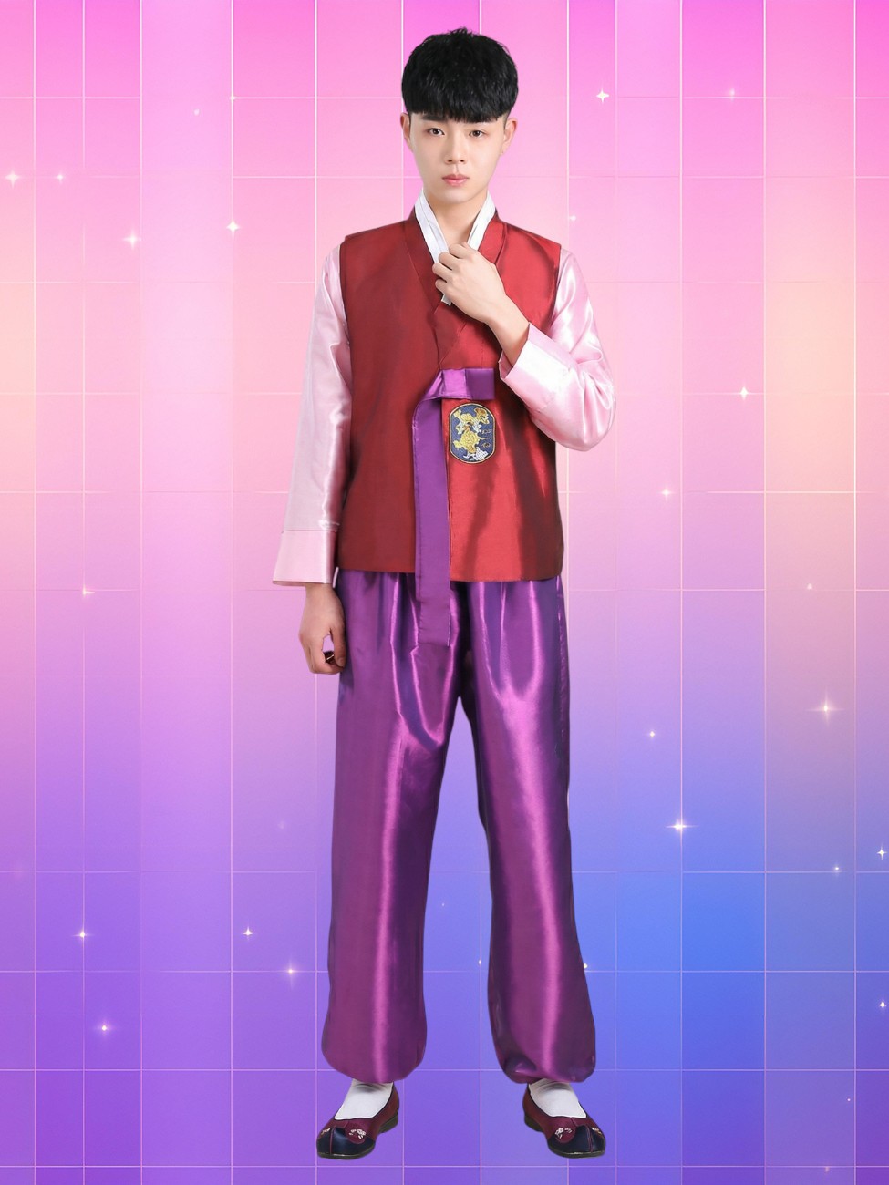 Un jeune homme asiatique porte le hanbok homme festif moderne couleurs vives avec veste rouge, pantalon violet brillant et chaussures traditionnelles, sur fond rose et bleu scintillant.