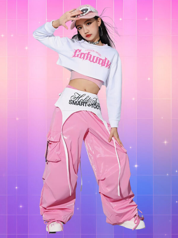 Une jeune fille portant une tenue kpop enfant streetwear rose 3 pièces avec sweat court blanc, top rose et pantalon large brillant, sur un fond rose et violet.