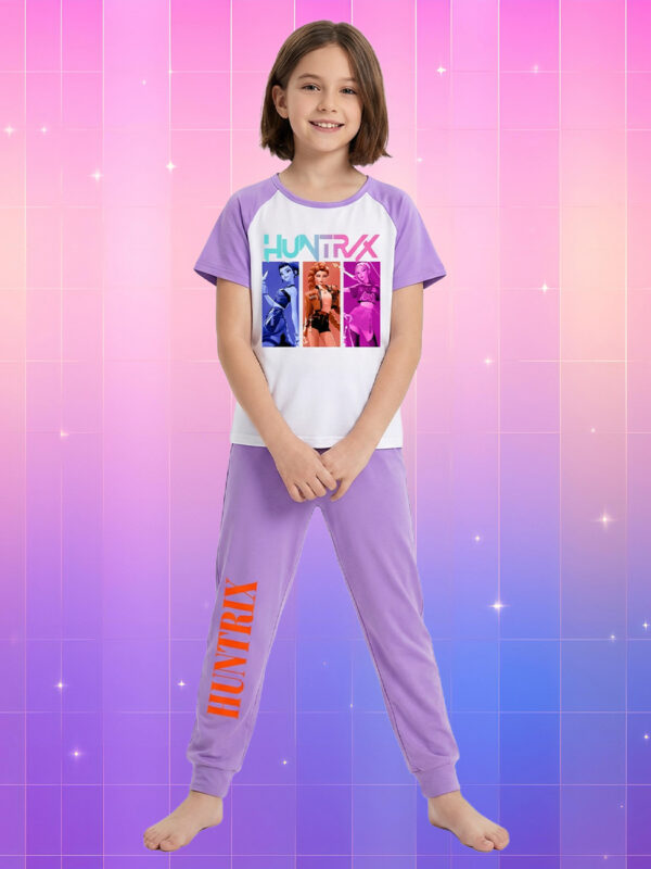 Une jeune fille souriante portant une tenue kpop enfant pyjama à manches courtes Kpop Demon Hunters, composée d'un t-shirt blanc et violet avec un imprimé graphique et d'un pantalon violet assorti, sur un fond quadrillé aux dégradés de rose et de violet parsemé d'étoiles.