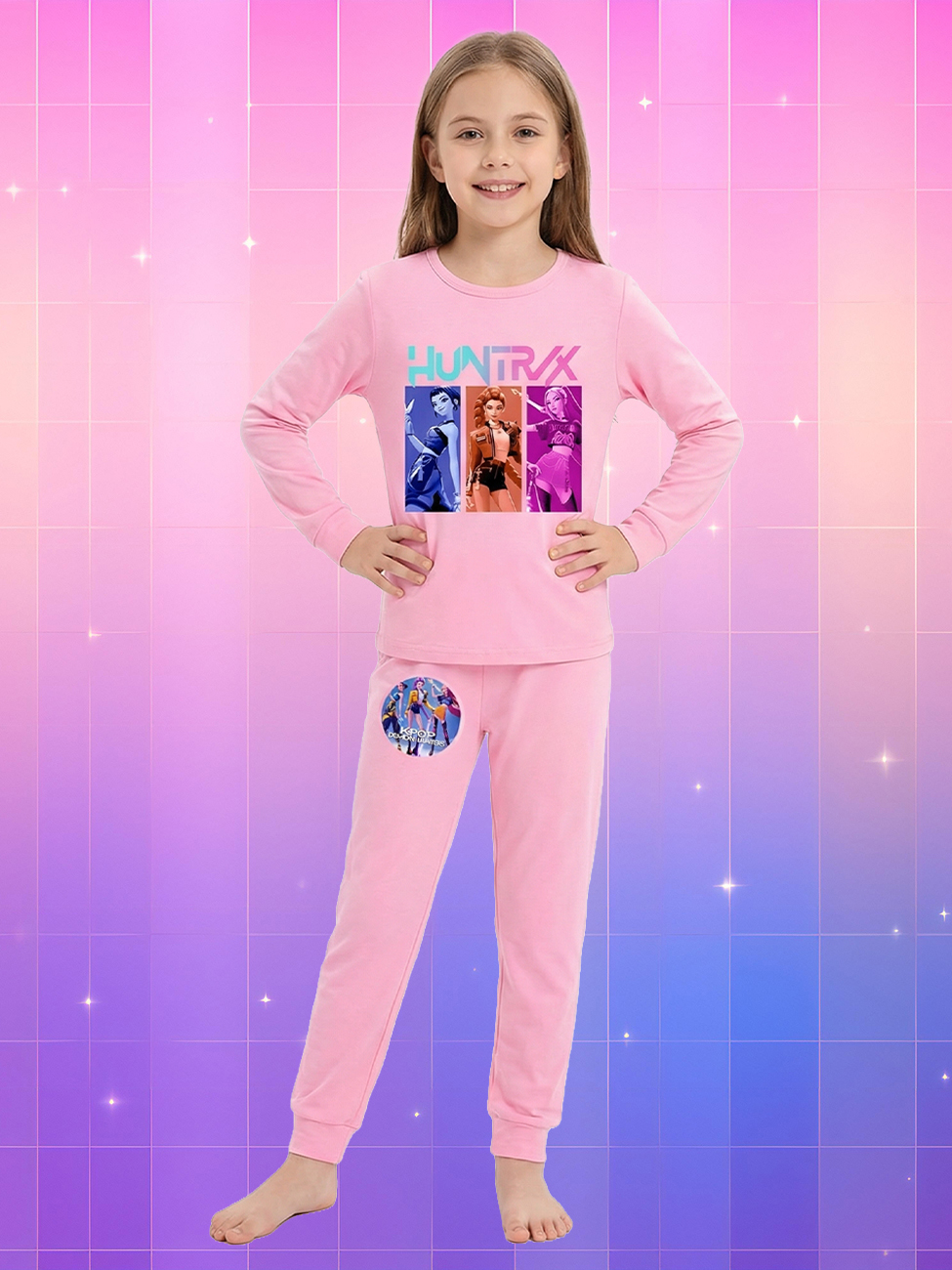 Une jeune fille souriante portant une tenue kpop enfant pyjama Kpop Demon Hunters rose à manches longues avec des motifs de personnages colorés, posant devant un background quadrillé aux nuances de rose, violet et bleu.