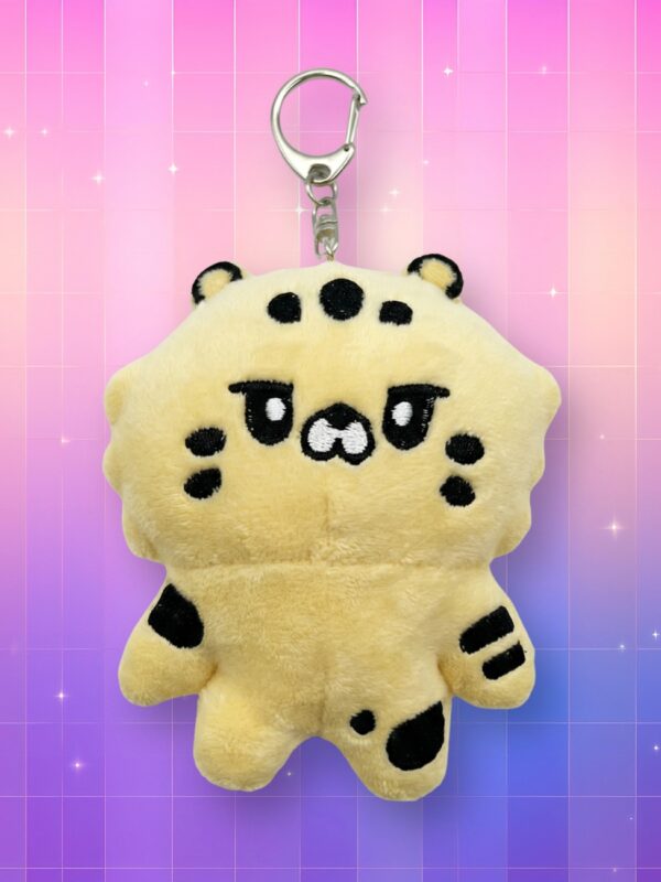 Une mignonne Peluche porte-clés Le Sserafim - Kimachi en forme de petit tigre jaune tout doux avec des taches noires et un crochet en métal argenté, présentée sur un fond quadrillé aux couleurs dégradées de rose et de violet parsemé de petites étoiles étincelantes.