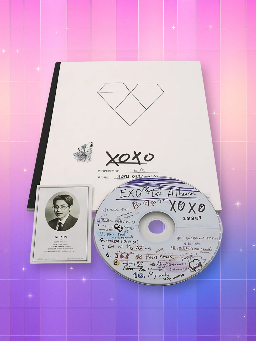L'album Exo 1er album Xoxo version Kiss présenté avec son CD illustré de dessins et sa carte de collection, posés sur un fond dégradé rose et violet scintillant.