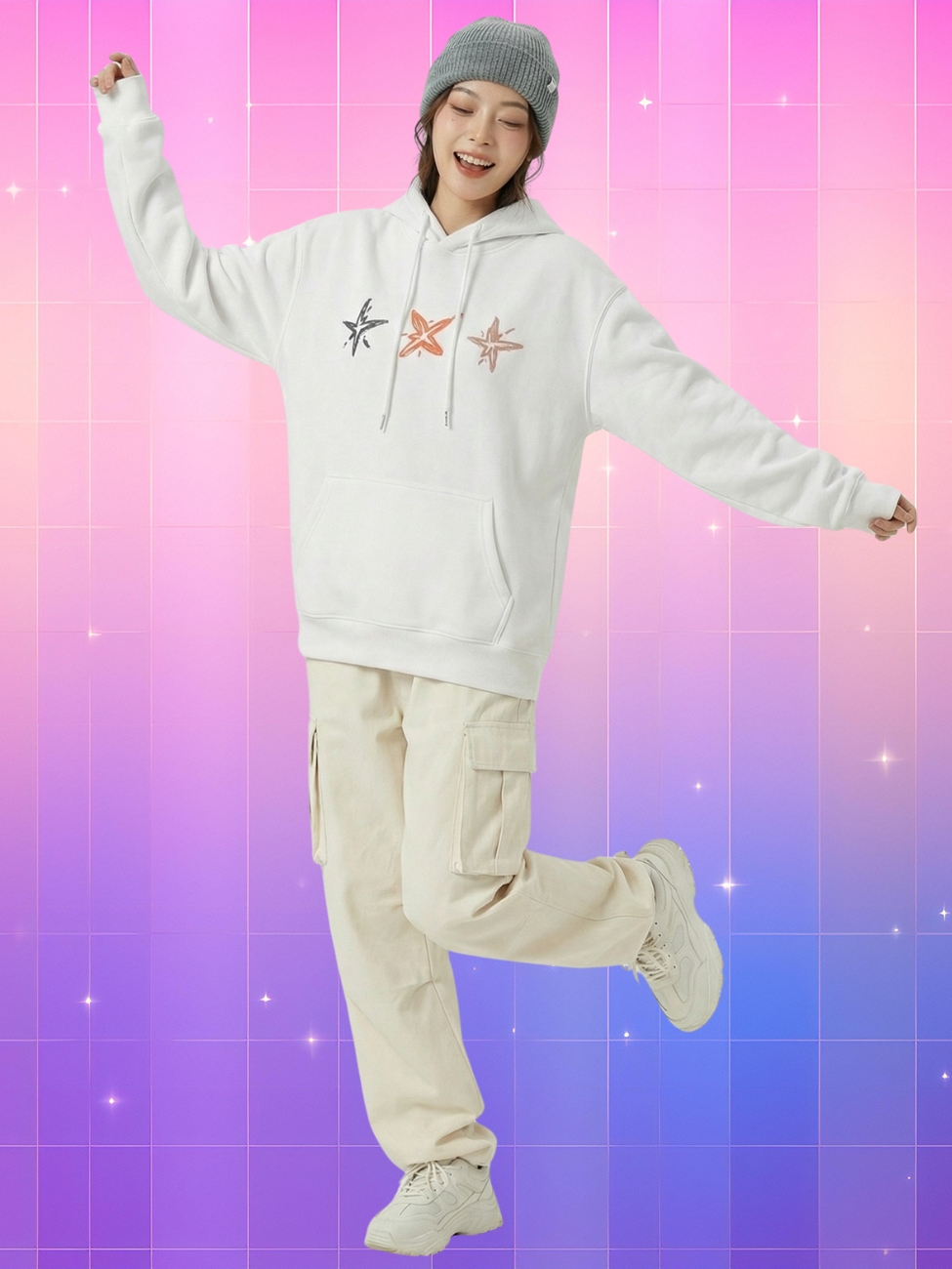 Une jeune femme souriante portant un sweat à capuche kpop TXT de couleur blanche avec trois étoiles dessinées sur la poitrine, posant joyeusement devant un arrière-plan quadrillé aux nuances de rose, violet et bleu.