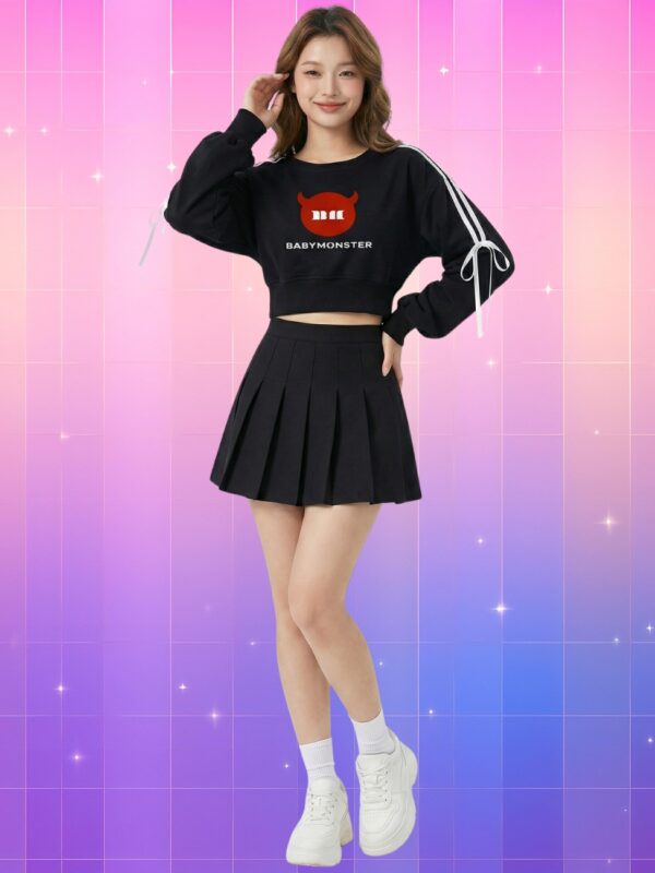 Une jeune femme souriante portant un Pull kpop crop-top Babymonster noir avec un logo rouge, une jupe plissée assortie et des baskets blanches, devant un fond dégradé rose et violet scintillant.