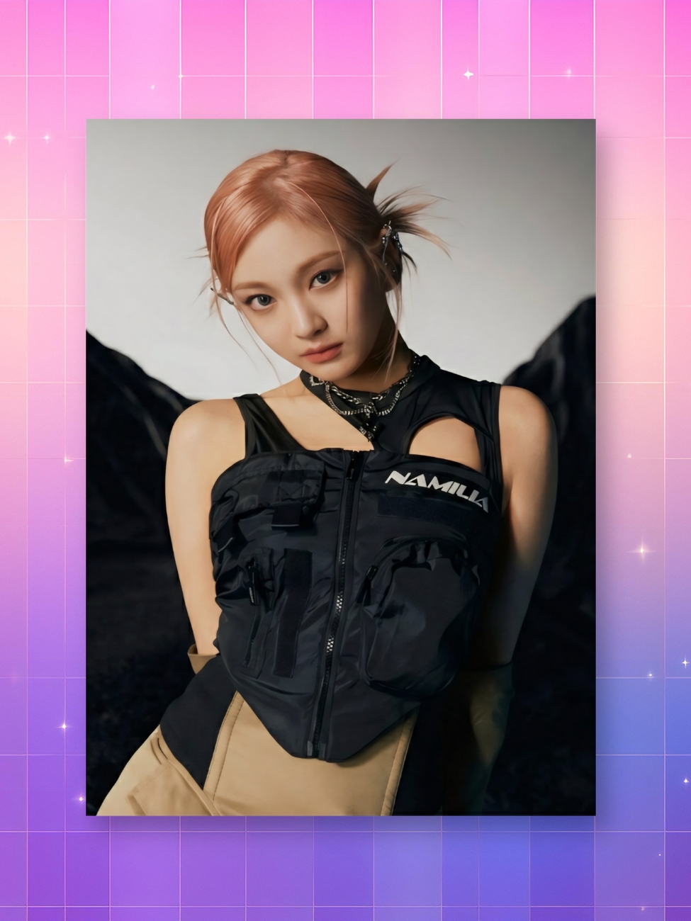 Un poster kpop Ningning en tenue Namilia montrant la chanteuse aux cheveux roses vêtue d'un haut noir technique avec des poches, posant devant un décor sombre de montagnes, le tout présenté sur un fond quadrillé rose et violet scintillant.
