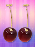 Des boucles d'oreille tendance en forme de cerise rouges et brillantes, suspendues à de longues tiges dorées sur un fond rose et violet scintillant.