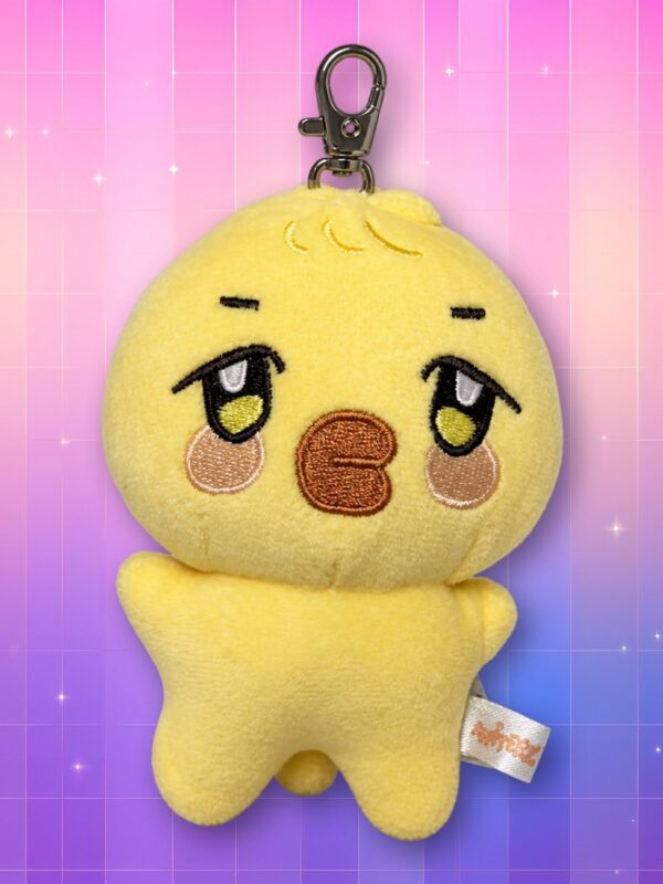Une peluche jaune en forme de petit personnage, appelée peluche porte-clés bbyongMING ANITEEZ, avec un mousqueton métallique, sur un fond rose et violet scintillant.