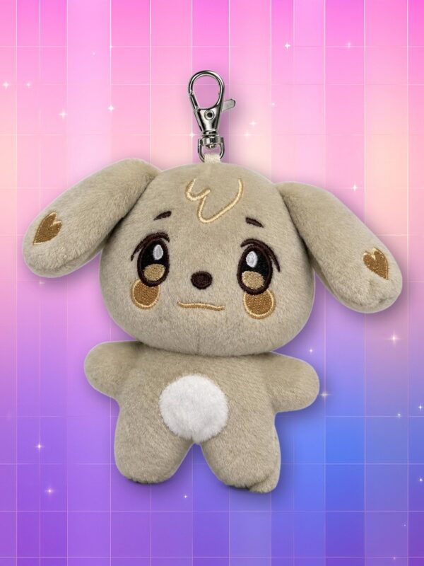 Une peluche porte-clés TYUdeongi ANITEEZ représentant un petit chien beige aux longues oreilles, avec un mousqueton argenté, sur un fond rose et violet scintillant.